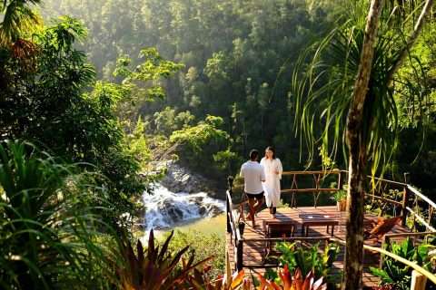 A couple relaxing on a wooden deck overlooking a cascading waterfall in a lush tropical jungle.