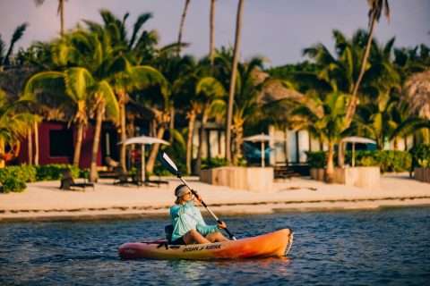 A person paddling an orange kayak in the water with a tropical beach resort and palm trees in the background.
