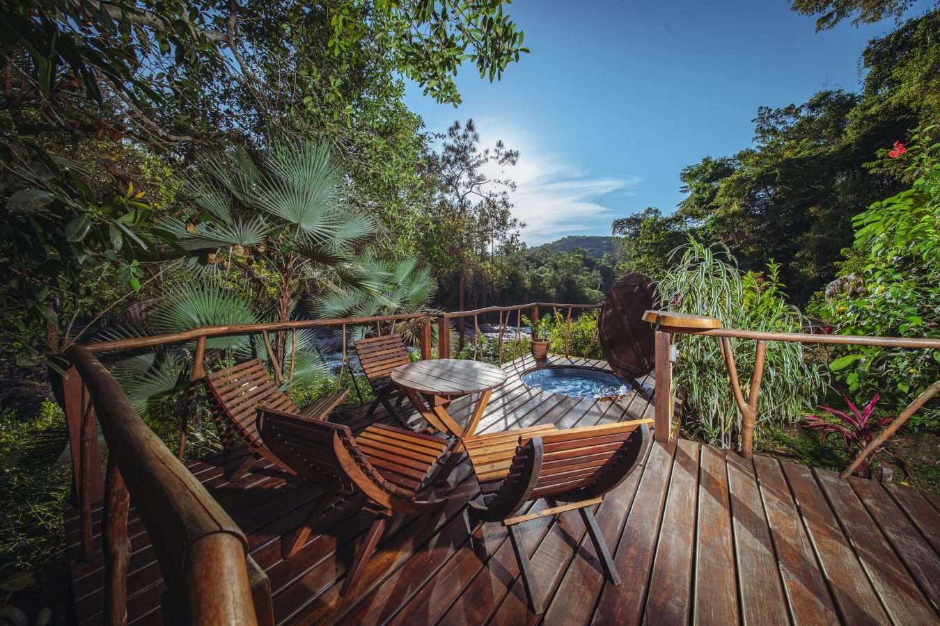 Private outdoor deck with a hot tub, round wooden table, and lounge chairs overlooking a rushing forest river.