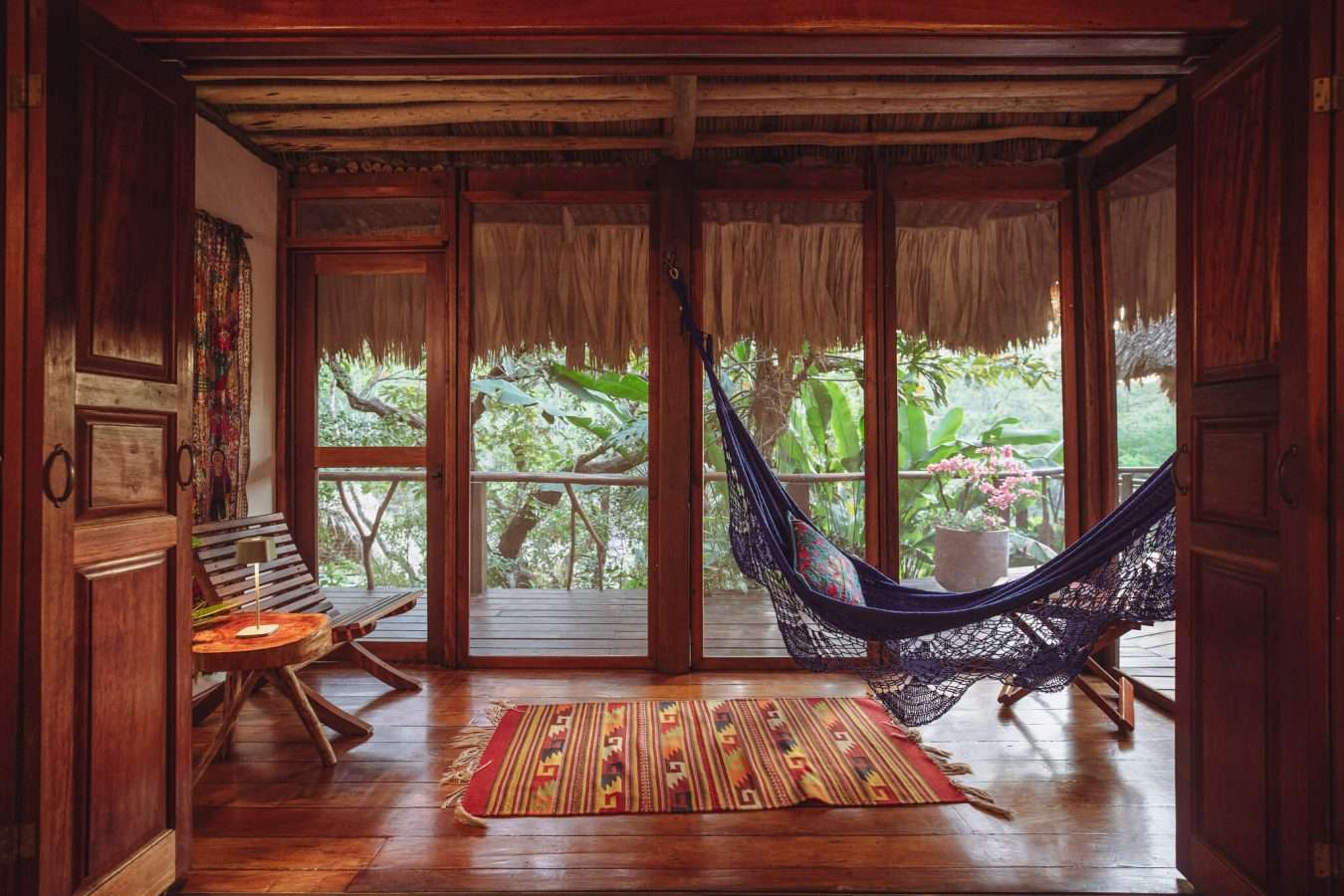 Interior lounge area with a woven hammock, patterned kilim rug, dark wood floors, and open doors leading to a private deck.