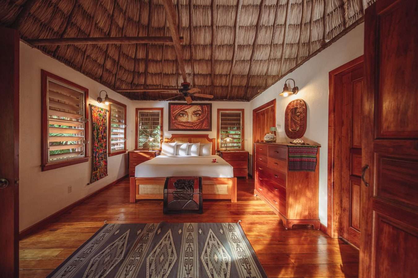 Bedroom with a king-sized bed, thatched vaulted ceiling with fan, Guatemalan textile accents, and jungle views through louvered windows.