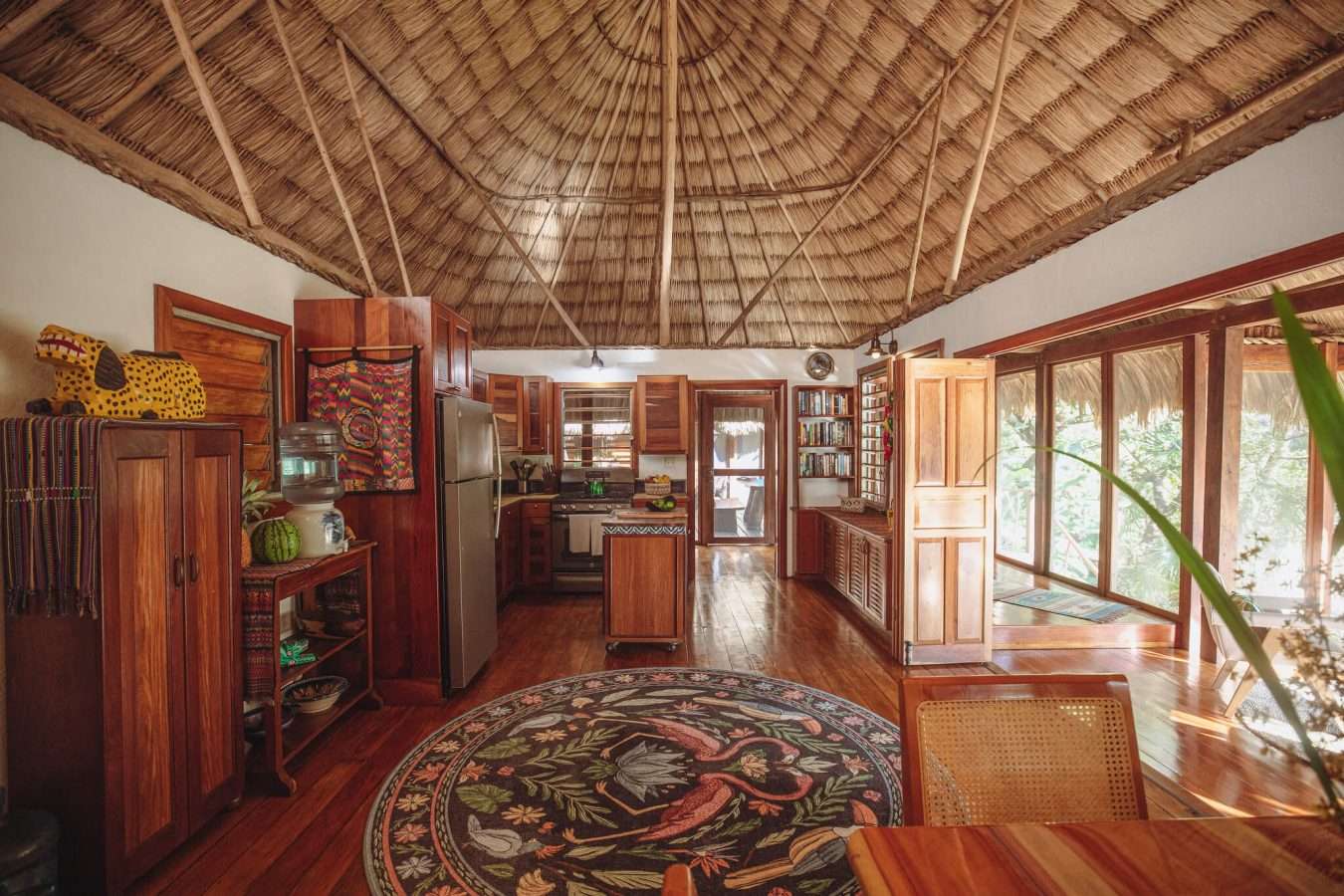 Open-plan kitchen and living area with a domed thatched ceiling, wood cabinetry, and screened doors opening to a jungle-view porch.